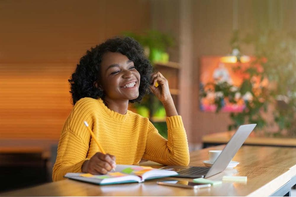 Young girl in yellow holding a notebook and pencil and looking at a laptop